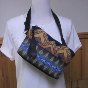 May include: A crossbody bag with a geometric pattern in shades of navy, yellow, and green. The bag has a black adjustable strap and a small black tassel. The bag is worn over the shoulder.