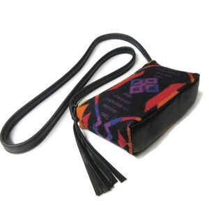 May include: A small, black crossbody bag with a geometric pattern in red, orange, purple, and blue. The bag has a black leather base, strap, and tassel. The strap is long and thin, and the tassel hangs from the side of the bag.