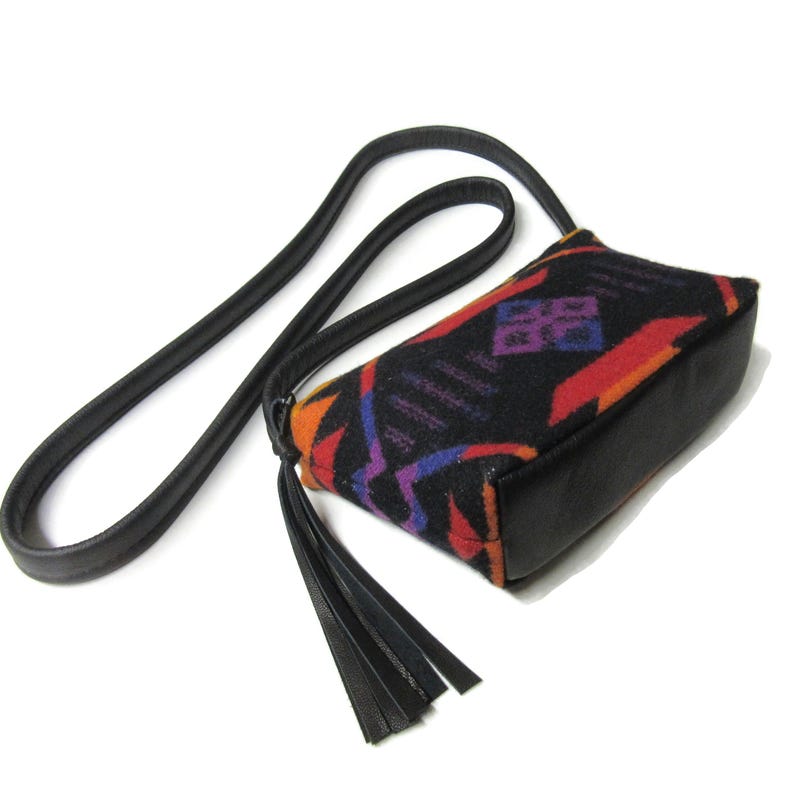 May include: A small, black crossbody bag with a geometric pattern in red, orange, purple, and blue. The bag has a black leather base, strap, and tassel. The strap is long and thin, and the tassel hangs from the side of the bag.