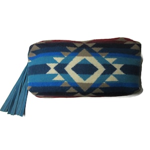 May include: A rectangular clutch with a geometric pattern in shades of blue, gray, and cream. The clutch has a blue leather fringe on one end. The design features diamond and triangle shapes.