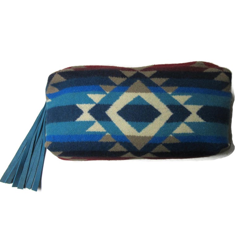 May include: A rectangular clutch with a geometric pattern in shades of blue, gray, and cream. The clutch has a blue leather fringe on one end. The design features diamond and triangle shapes.