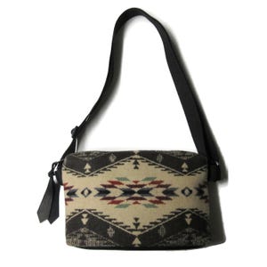 May include: A rectangular crossbody bag with a black strap and a patterned design. The bag features a cream-colored background with a geometric pattern in shades of brown, red, and blue. The strap is adjustable and made of black fabric.