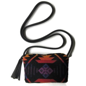 May include: A small, rectangular black crossbody bag with a colourful geometric pattern in orange, red, purple, and blue. The bag has a black strap and a black tassel. The bag is approximately 20 cm wide.