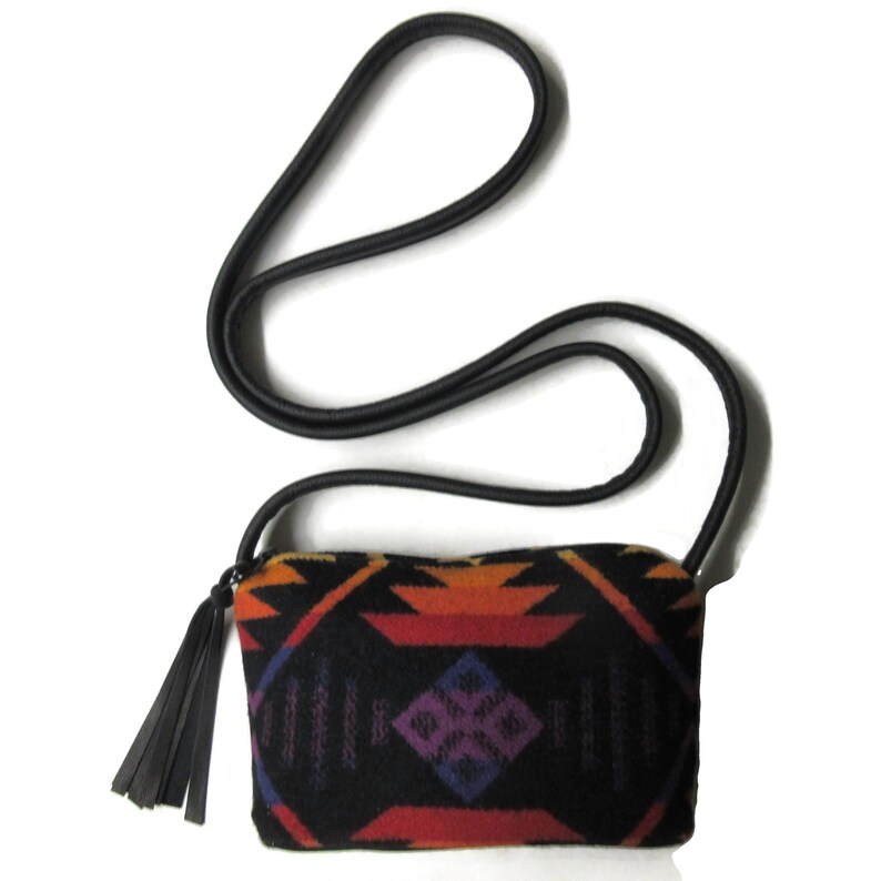 May include: A small, rectangular black crossbody bag with a colourful geometric pattern in orange, red, purple, and blue. The bag has a black strap and a black tassel. The bag is approximately 20 cm wide.