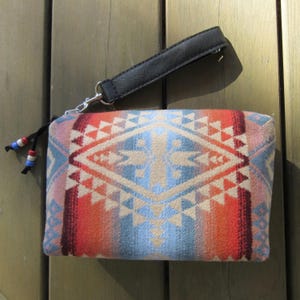 May include: A rectangular clutch with a black leather wrist strap and a zipper closure. The clutch features a geometric pattern in shades of blue, orange, red, and beige. Small beads in red, white, and blue adorn the zipper pull.