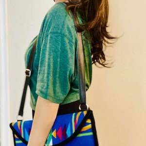 May include: A blue and black shoulder bag with a colorful geometric pattern. The bag has a black strap and trim. The pattern includes red, yellow, and turquoise shapes. The bag is being worn over the shoulder.