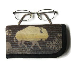 May include: A pair of thin-rimmed metal eyeglasses with oval lenses positioned above a rectangular fabric eyeglass case. The case features a woven pattern of a large buffalo in shades of tan and brown, finished with a solid black fabric trim.