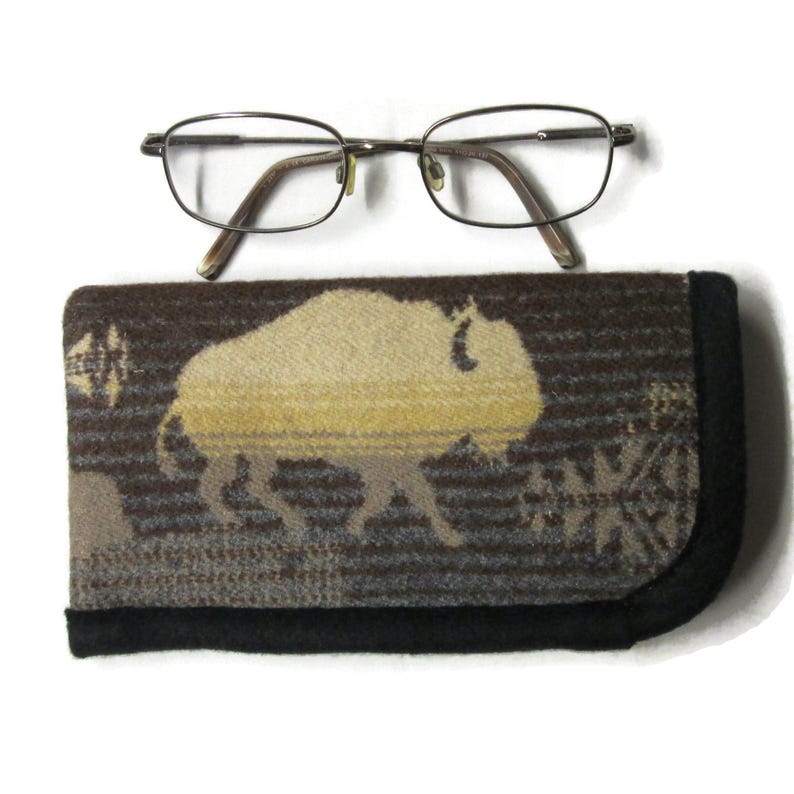 May include: A pair of thin-rimmed metal eyeglasses with oval lenses positioned above a rectangular fabric eyeglass case. The case features a woven pattern of a large buffalo in shades of tan and brown, finished with a solid black fabric trim.