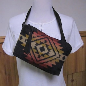 May include: A black shoulder bag with a geometric pattern in shades of red, yellow, and beige. The bag has a long, black strap and a zipper closure. The pattern is a repeating diamond design.