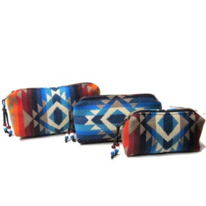 May include: Three zippered pouches with a geometric pattern in shades of blue, red, orange, and gray. Each pouch has a black zipper and a beaded tassel. The pouches vary in size, with the largest approximately 9 inches long.