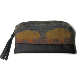 May include: A rectangular clutch with a black leather bottom and a gray fabric top featuring two brown bison silhouettes. The clutch has a black zipper and a black tassel attached to the side. The design is likely inspired by nature and wildlife.
