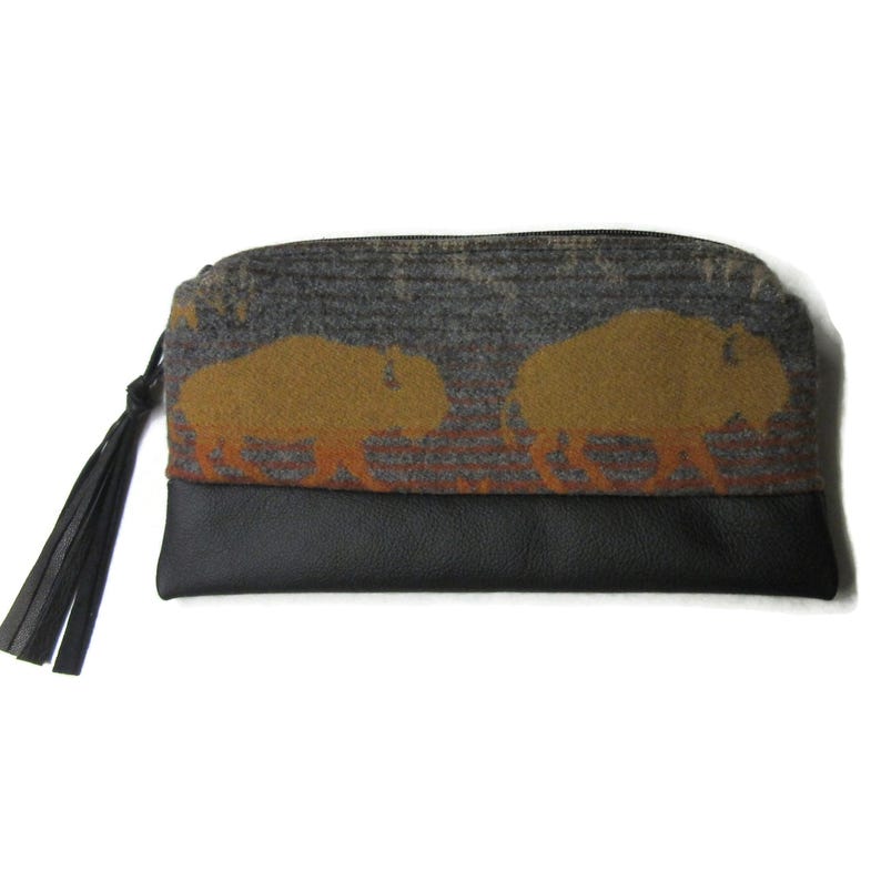 May include: A rectangular clutch with a black leather bottom and a gray fabric top featuring two brown bison silhouettes. The clutch has a black zipper and a black tassel attached to the side. The design is likely inspired by nature and wildlife.