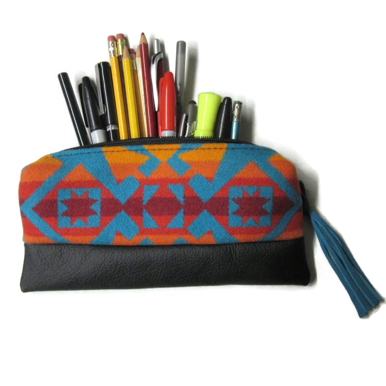 K&ouml;nnte beinhalten: Ein Federm&auml;ppchen mit einem s&uuml;dwestlichen Muster in Orange, Blau und Rot und einem schwarzen Lederboden. Das Etui ist mit Stiften, Bleistiften und Markern gef&uuml;llt. Eine blaue Quaste h&auml;ngt am Rei&szlig;verschluss.