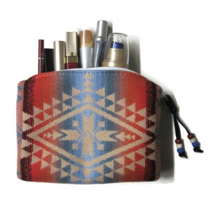 May include: A patterned cosmetic bag in shades of red, blue, and beige, filled with various makeup items. The bag features a geometric design and a black drawstring with decorative beads. The bag is designed to hold makeup.