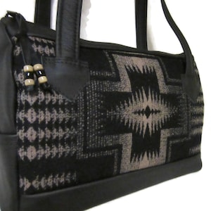 May include: A black handbag featuring a central geometric pattern in shades of black and grey. The bag includes black leather-like trim, two sturdy handles, and a zipper closure adorned with two small, light-colored cylindrical beads on a cord.