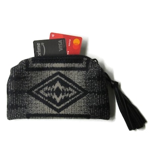 May include: A small, patterned wallet in shades of gray and black, with a diamond design in the center. The wallet has a black zipper and a black tassel. Two credit cards are visible inside the wallet.