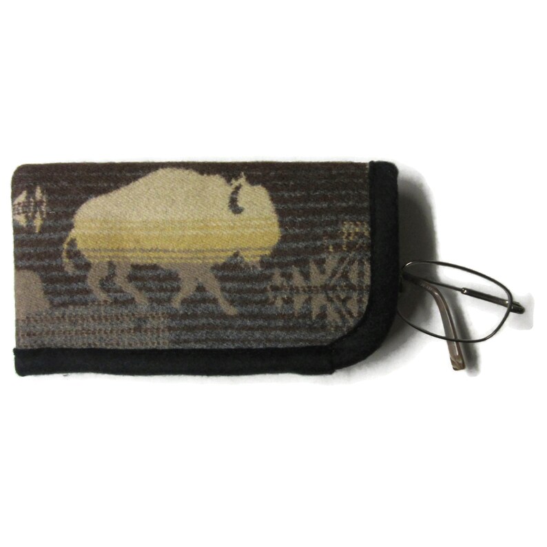 May include: A rectangular eyeglass case featuring a woven pattern of a light-colored buffalo against a dark brown and gray striped background. The case has a solid black fabric trim along the edges and is shown with a pair of thin-framed glasses.