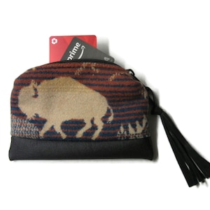 May include: A small, patterned coin purse with a bison design. The purse features a black leather bottom and a zipper closure with a black tassel. Two cards are visible inside the purse.