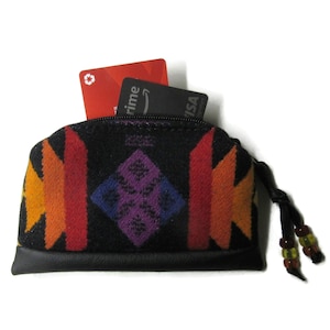 May include: A small, black zippered wallet with a colorful geometric pattern in orange, red, and yellow. The wallet has a black leather base and a black tassel with beads. Two credit cards are visible inside.