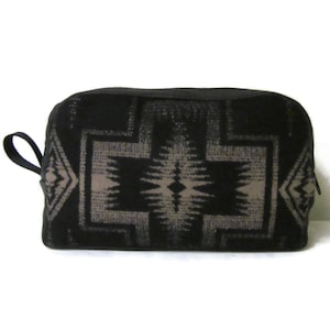 May include: A rectangular zippered pouch featuring a black and grey geometric pattern. The design consists of a large central diamond-shaped motif flanked by smaller diamond shapes on the sides. A black fabric loop is attached to the left side.
