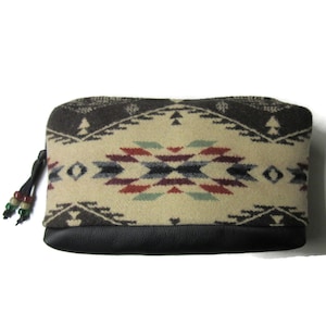 Op de afbeelding: Een rechthoekig etui met een cr&egrave;me, bruin, rood en blauw Southwestern patroon. Het bovenste gedeelte heeft een geometrisch ontwerp, terwijl de onderkant zwart is. Een zwarte ritstrekker met decoratieve kralen bevindt zich aan de linkerkant.