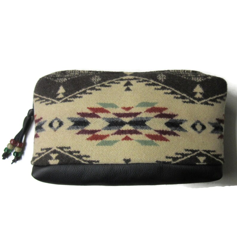 Op de afbeelding: Een rechthoekig etui met een cr&egrave;me, bruin, rood en blauw Southwestern patroon. Het bovenste gedeelte heeft een geometrisch ontwerp, terwijl de onderkant zwart is. Een zwarte ritstrekker met decoratieve kralen bevindt zich aan de linkerkant.