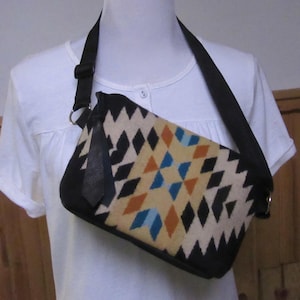 May include: A rectangular crossbody bag with a black strap and geometric pattern. The bag features a cream-colored background with black, orange, and blue diamond shapes. The bag has a black leather accent and a black strap.