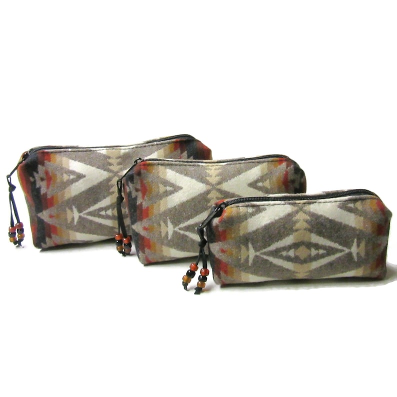 May include: Three zippered pouches with a geometric pattern in shades of brown, white, orange, and red. The pouches have black zipper pulls and black tassels with brown beads.