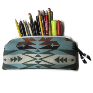 May include: A turquoise pencil case with a Southwestern pattern, filled with various pens and pencils. The case has a zipper closure and a black cord with decorative beads. The pencils and pens are of different colors and sizes.