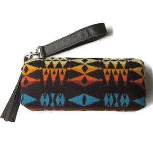 May include: A rectangular clutch with a geometric pattern in shades of brown, orange, yellow, and blue. It has a dark brown leather wrist strap and a tassel. The clutch is designed with a zipper closure.