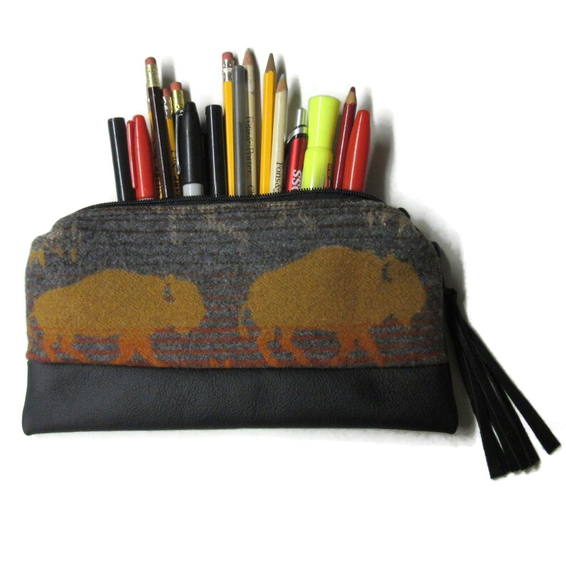 May include: A gray and black pencil case with a bison design. The case is filled with pencils, pens, and markers. The bison are in a golden-yellow color, and the bottom of the case is black. A black tassel hangs from the side.