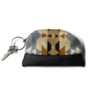 May include: A small key chain with a black leather bottom and a top made of patterned fabric in shades of brown, gray, and white. A silver key is attached to the key ring.
