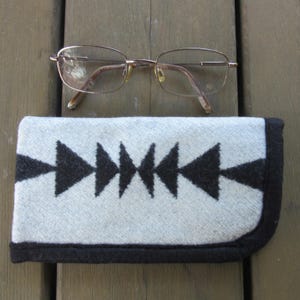 May include: Eyeglasses with a gold-colored frame rest above a gray and black patterned glasses case. The case features a geometric design of black triangles on a light gray background, with a black border.