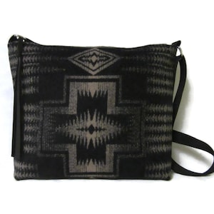 May include: Black and gray crossbody bag with a geometric pattern. The bag features a long, adjustable strap and a black leather tassel. The bag's design includes diamond and cross-like shapes, creating a Southwestern or Native American aesthetic.