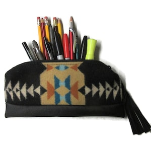 May include: A black pencil case with a geometric pattern in beige, orange, and blue. The case is filled with various pens and pencils. The bottom of the case is black, and it has a black tassel attached to the zipper.
