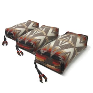 May include: Three rectangular patterned fabric pouches with black zipper closures and brown leather tassels with wooden beads. The pouches are made of a soft, woven fabric with a geometric design in shades of brown, white, red, and black.