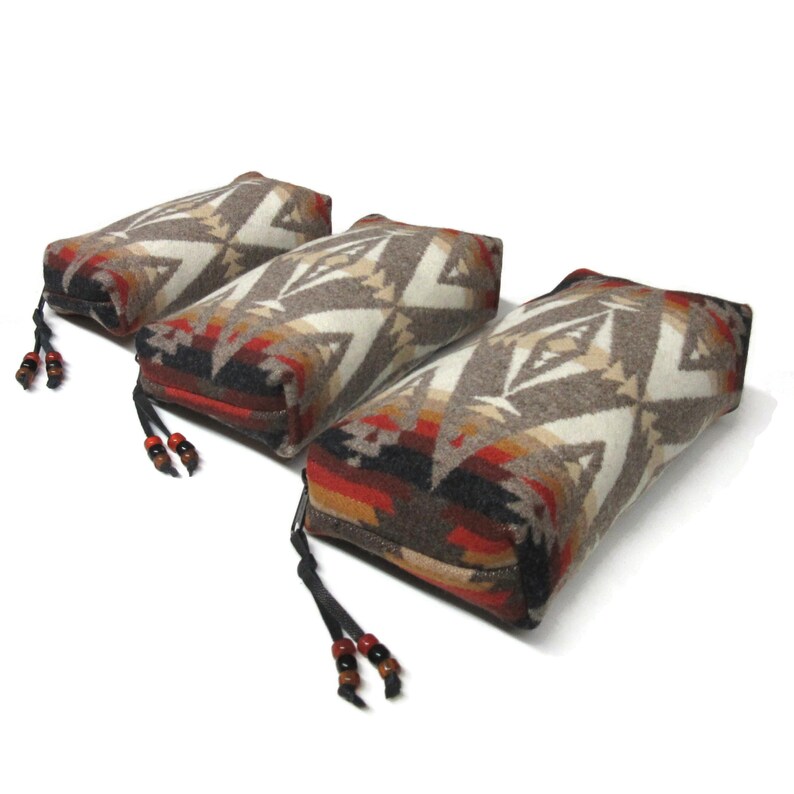 May include: Three rectangular patterned fabric pouches with black zipper closures and brown leather tassels with wooden beads. The pouches are made of a soft, woven fabric with a geometric design in shades of brown, white, red, and black.
