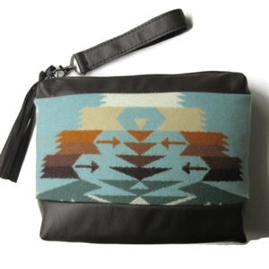 May include: A rectangular clutch with a brown leather base and top, and a patterned fabric center. The fabric features a geometric design in shades of blue, brown, and beige. A matching leather wrist strap and tassel are attached.