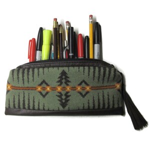 May include: A rectangular pencil case made of sage green fabric with a dark brown leather base and a matching leather zipper pull. The fabric features a geometric pattern in black, orange, and yellow. It is filled with various pens and pencils.