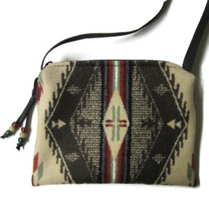 May include: A rectangular beige crossbody bag with a geometric pattern in shades of brown, red, and green. The bag has a black strap and a zipper closure. The zipper pull has decorative beads.