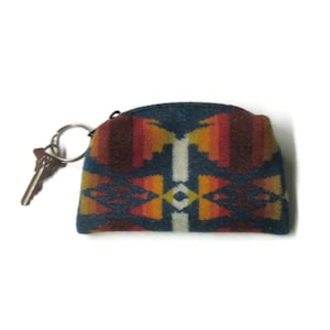 May include: A small, blue key pouch with a Southwestern-inspired geometric pattern in shades of red, orange, yellow, and white. A silver key ring holds a single key. The pouch is made of a textured fabric.
