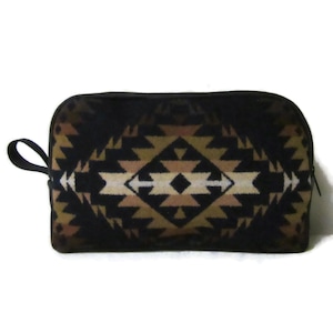 May include: A rectangular zippered pouch featuring a geometric pattern in shades of black, tan, and cream. The bag has a rounded top edge, a black fabric wrist strap on the left side, and a black zipper closure along the top and side edges.