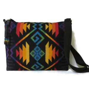 May include: A black crossbody bag with a vibrant geometric pattern in rainbow colors. The bag features a long black strap and a small beaded detail on the side. The design includes orange, yellow, green, blue, and purple shapes.