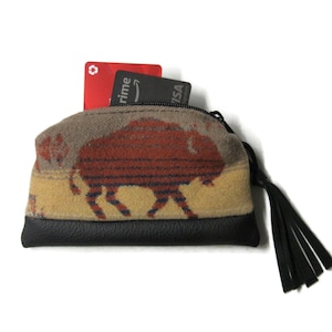 May include: A small, zippered coin purse with a bison design. The purse features a black leather bottom, a tan and brown patterned body, and a black tassel. Two credit cards are visible inside the purse.