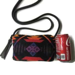 May include: A small, rectangular black crossbody bag with a colourful geometric pattern in orange, red, purple, and blue. The bag has a black strap and a black tassel. A red Coca-Cola can is next to the bag.