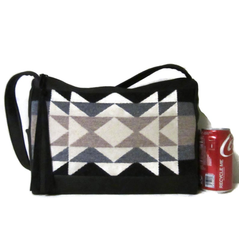 May include: Black shoulder bag with a geometric pattern in shades of gray, beige, and white. The bag has a black strap and a tassel on the side. The bag is approximately 12 inches wide.
