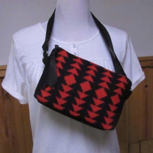 May include: Black and red geometric pattern crossbody bag with a black adjustable strap. The bag is rectangular and features a repeating pattern of red triangles on a black background. The strap is attached to the bag with silver-colored hardware.