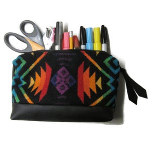 May include: A black pencil case with a colorful geometric pattern and a black leather bottom. The case is filled with various pens, pencils, scissors, and a stapler. The zipper has a black pull.