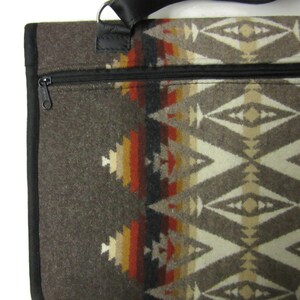 May include: A brown tote bag with a geometric pattern in shades of beige, red, and yellow. The bag has a black zipper, black trim, and a black strap. The bag is made of a thick, woven material.