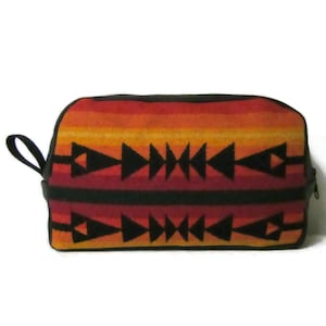 May include: A rectangular zippered pouch featuring a horizontal gradient pattern of red, orange, and yellow stripes. The pouch is decorated with two rows of black geometric arrow motifs and includes a black side handle and a black zipper closure.